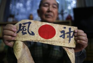 Senhor de meia idade segurando uma bandeira de cabeça, escrito ''kamikaze'', em japonês, e a bandeira do Japão entre as ''siglas'' japonesas.